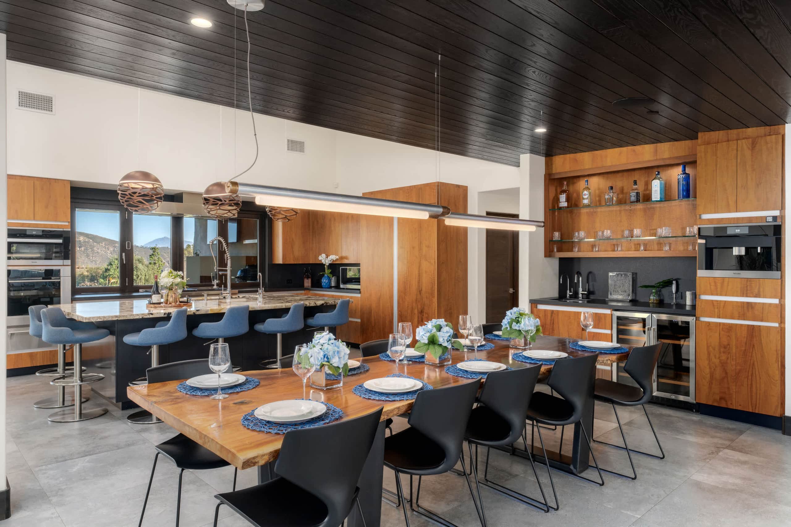 Long dining table set for a meal beside a kitchen bar with warm wood cabinetry